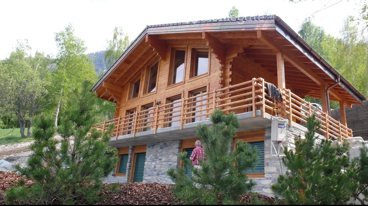 Two-story wooden house with a large balcony, stone base, and wooden upper level. Surrounded by trees and greenery.