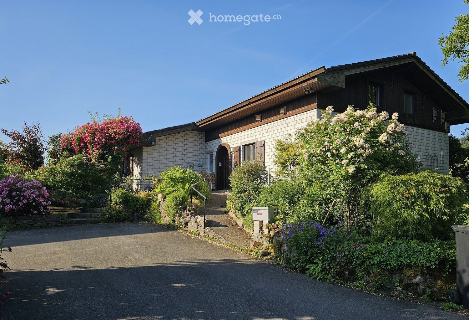 Two-story house with a tiled roof, white brick facade, and a well-maintained garden with colorful flowers and shrubs.