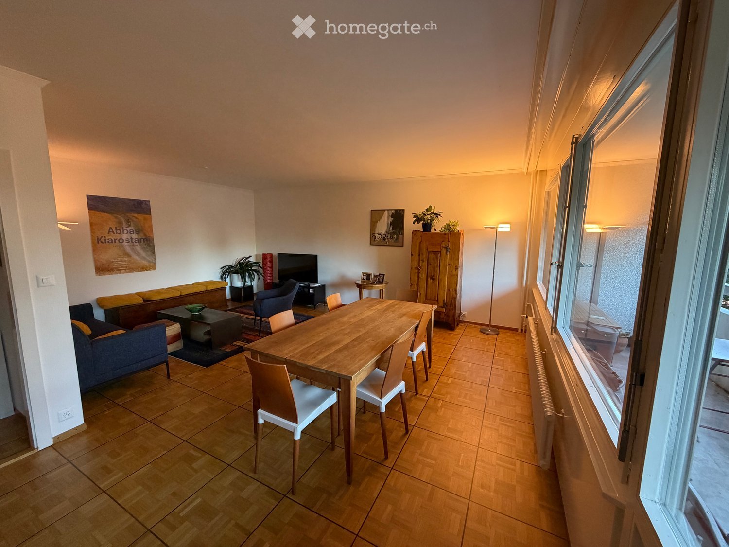 Bright, open living and dining area with wooden flooring, large windows, a sofa, coffee table, dining table with chairs, TV, and decorative items.