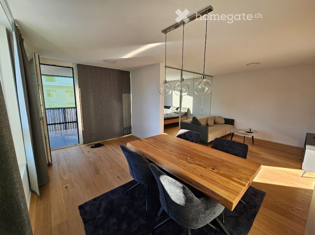 Open-plan living and dining area with wooden flooring, a large wooden dining table with chairs, a sofa, a mirror, and a bed visible in the background. Large windows and a sliding door lead to a balcony.