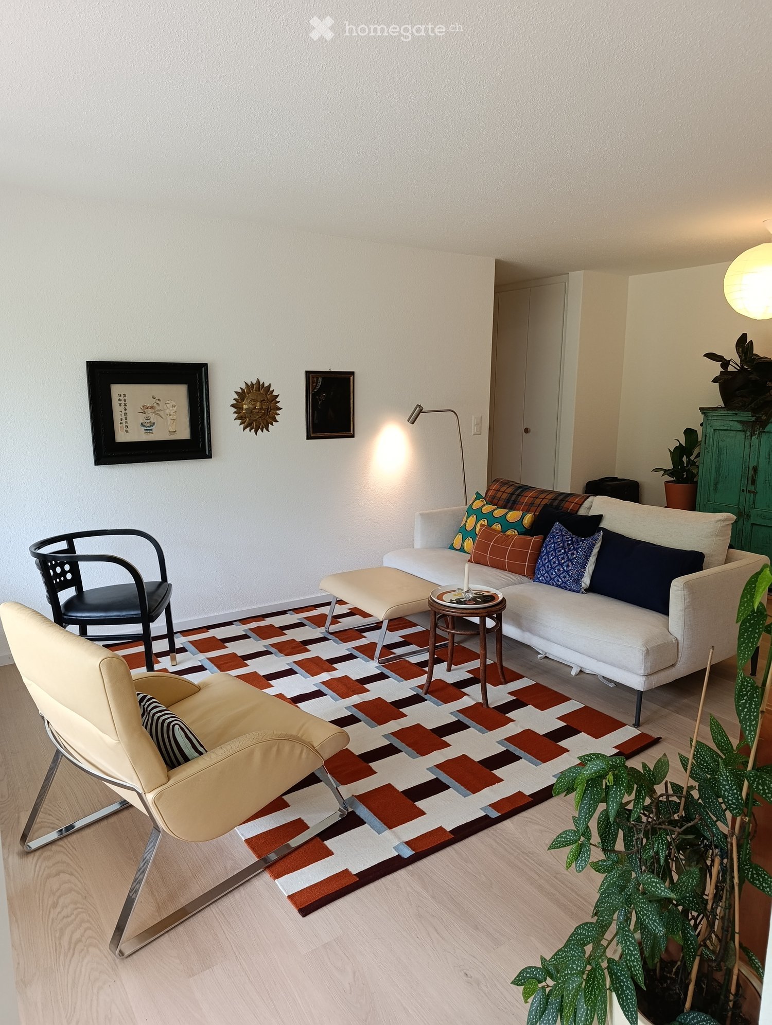 Modern living room with a white sofa, colorful cushions, a coffee table, a patterned rug, and a sleek chair. The room features wooden flooring, wall-mounted lighting, and decorative wall art.