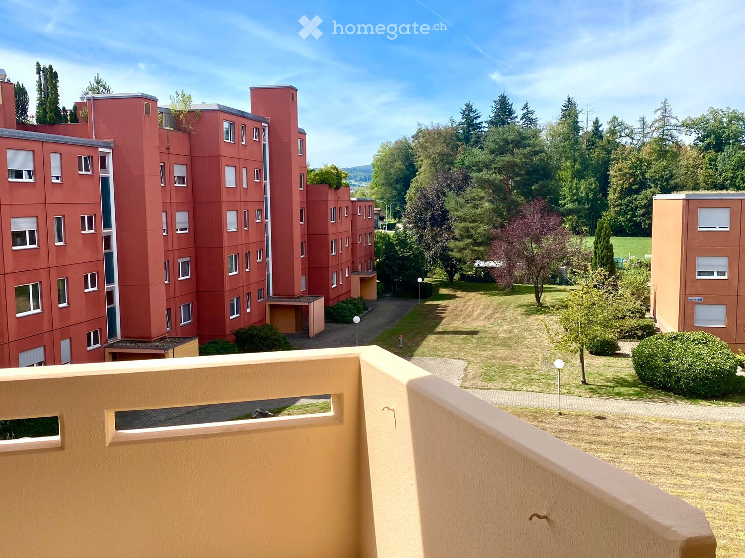 View from a balcony showing a multi-story red brick residential building surrounded by greenery and trees.