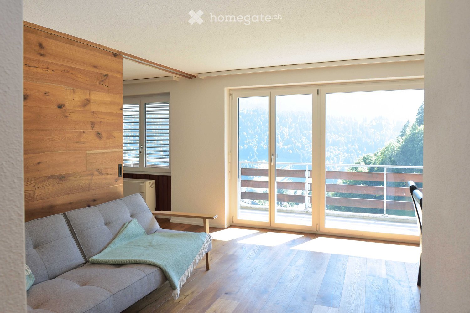 Bright living room with wooden flooring, grey sofa, throw blanket, small bench, large sliding glass doors leading to a balcony with mountain view.