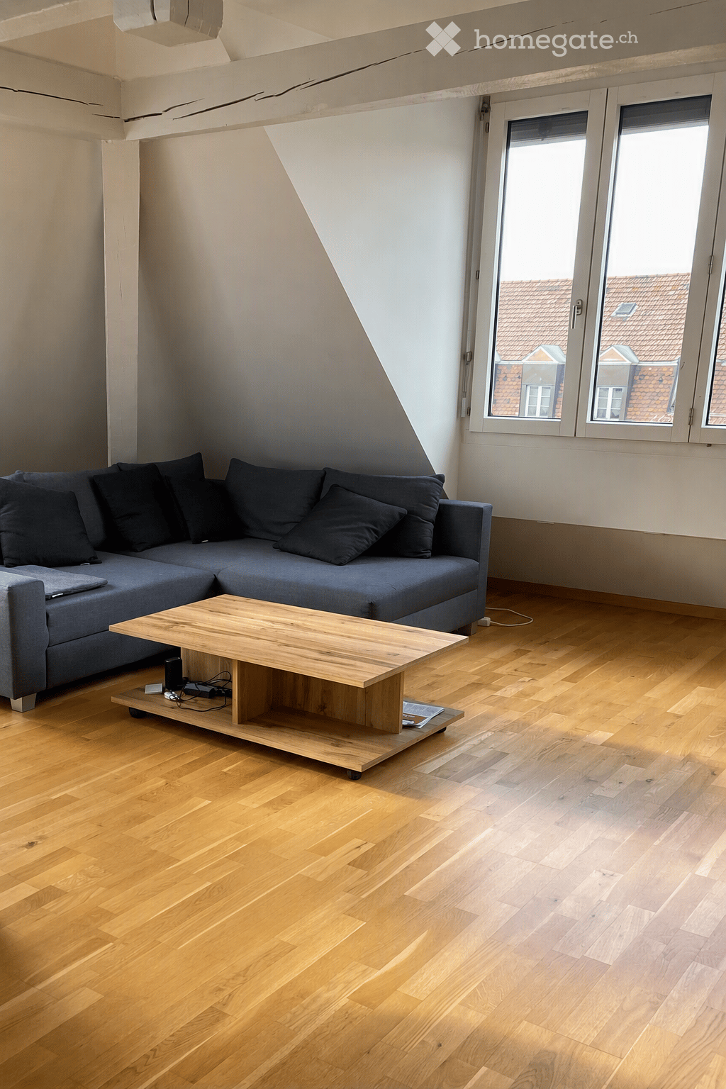 Modern living room with a grey sectional sofa, wooden coffee table, and hardwood flooring. Large windows offer a view of the city.