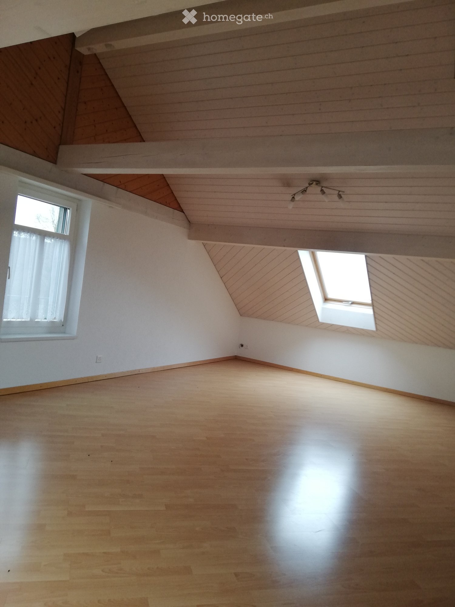 Empty room with wooden flooring, white walls, and a slanted wooden ceiling. Features include a large window, a skylight, and a track light fixture.