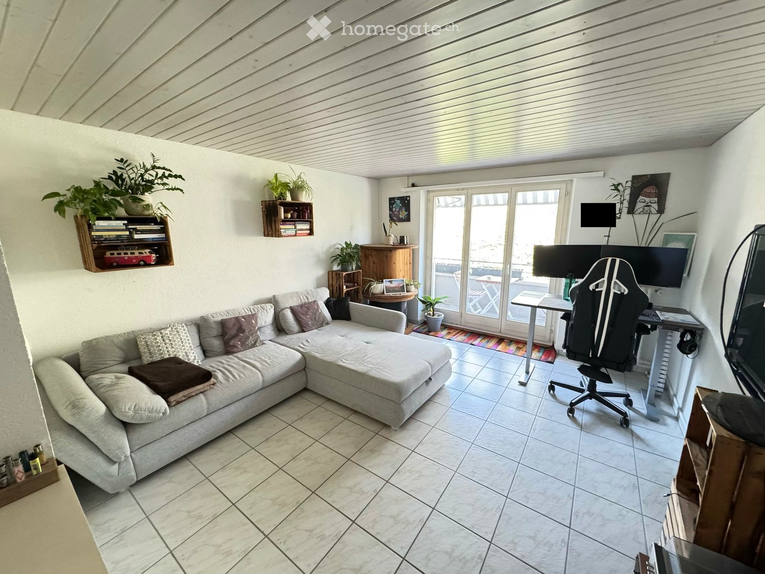 A modern living room with a grey sectional sofa, a black office chair, a small table, a TV screen, shelves with books and plants, and a large window leading to a balcony.
