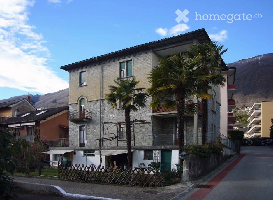 A multi-story stone building with balconies and palm trees in front, located in a residential area with a clear sky and distant mountains.