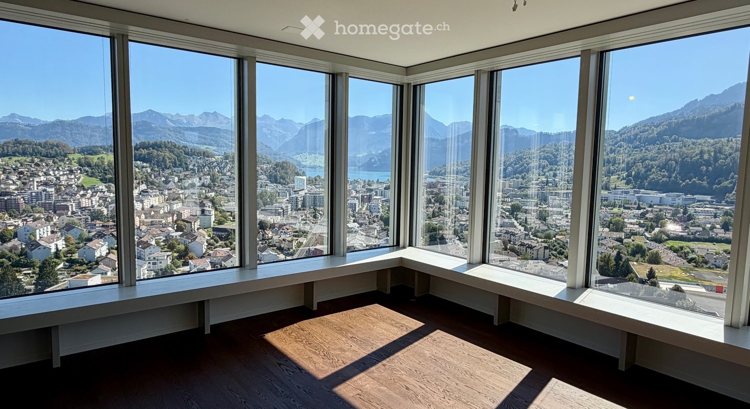 Empty room with large windows offering a panoramic view of a cityscape and mountains. The floor is wooden, and there is a white bench along the windowsill.