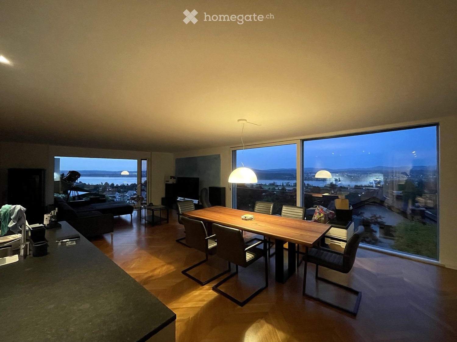 Open-plan living and dining area with large windows offering a scenic view, featuring a sofa, dining table with chairs, and a television.