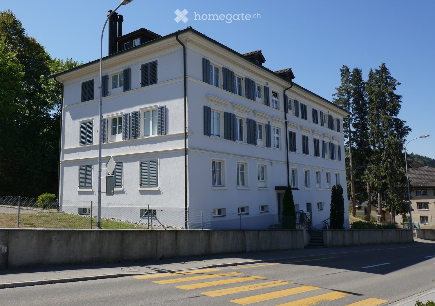 A large, white, multi-story building with numerous windows and blue shutters, situated on a raised ground floor with a concrete barrier and steps leading to the entrance.