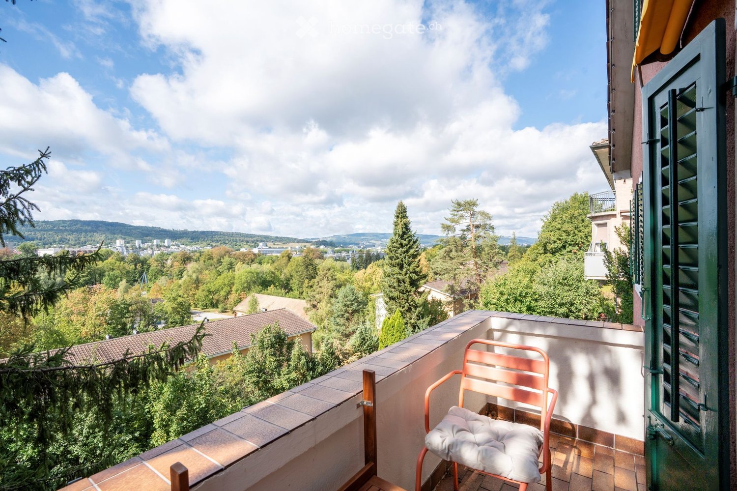 Balcony with a view of a cityscape and surrounding greenery, featuring a single chair with a cushion.