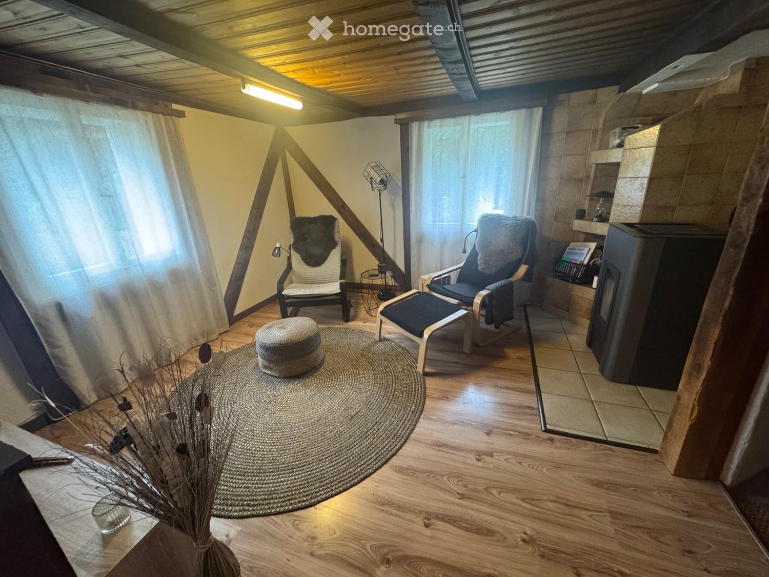 Cozy attic room with wooden floors, slanted ceilings, and large windows. Features two armchairs, a small round table, a fireplace, and a decorative plant arrangement.