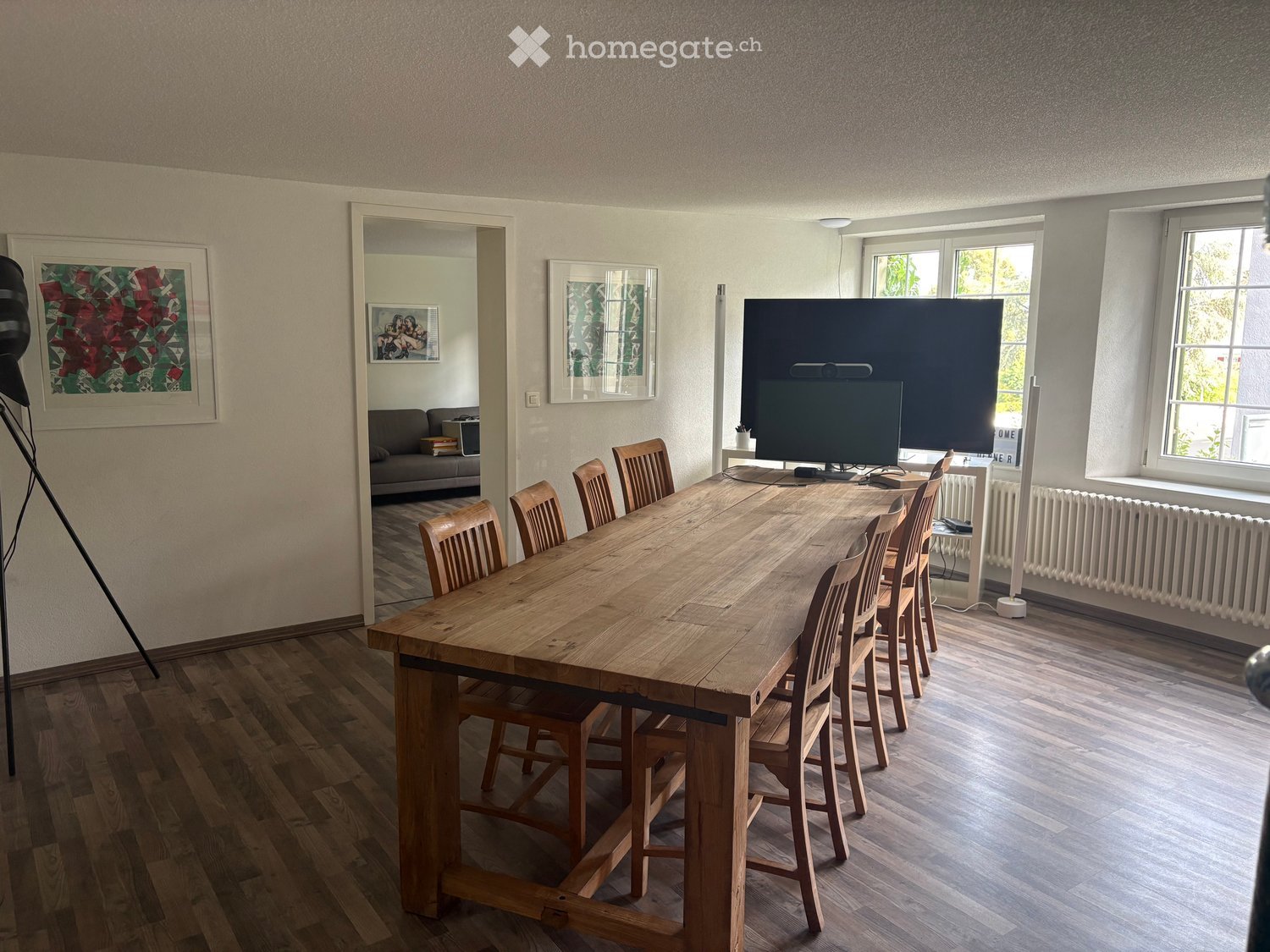 Dining room with a large wooden dining table surrounded by wooden chairs, a large screen, radiator, and picture frames on the walls.
