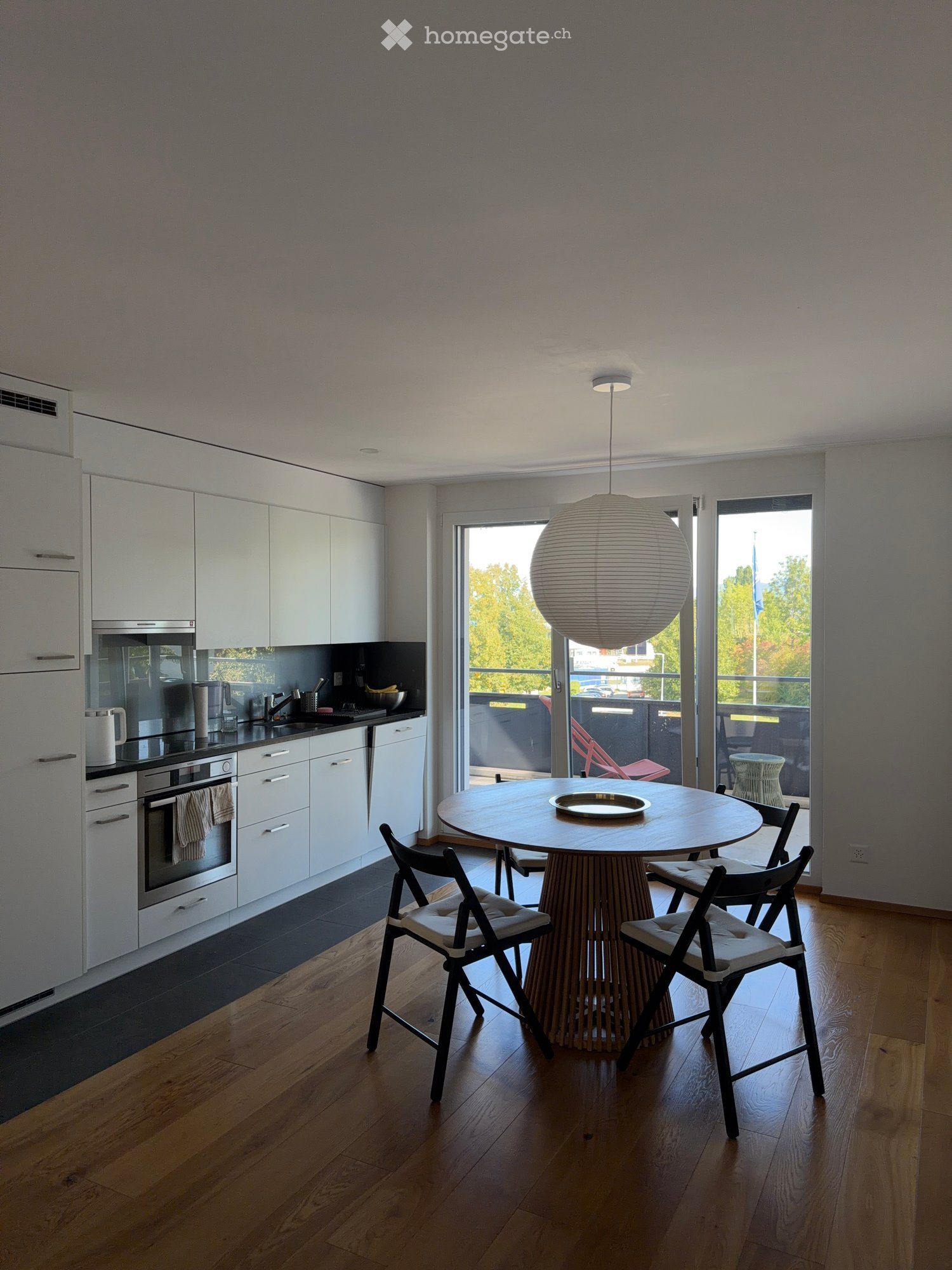 Modern kitchen with white cabinets, stainless steel appliances, a round wooden dining table with four chairs, and a hanging spherical lamp.