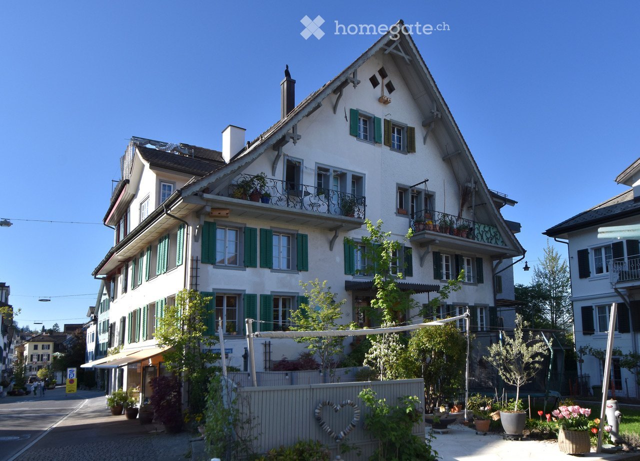 A multi-story residential building with white walls, green shutters, and balconies adorned with potted plants. The building features a classic architectural style with a pitched roof and multiple windows.