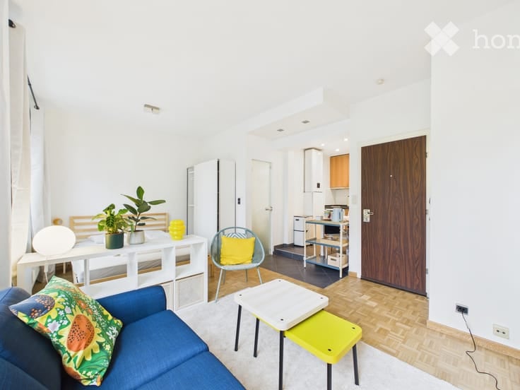 Bright, modern living space with a blue sofa, white coffee table, yellow accent chair, small kitchen area, and decorative plants.