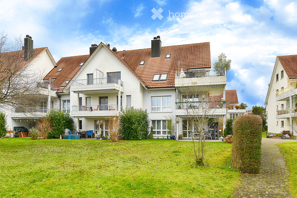 Two-story white house with red-tiled roof, multiple balconies, and surrounding greenery.
