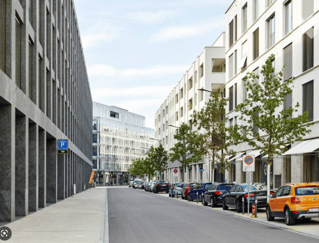 Urban street lined with modern buildings, parked cars, and trees. The buildings are contemporary with clean lines and glass facades.
