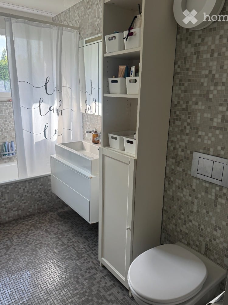 Modern bathroom with a white toilet, sink, and built-in cabinet with shelves and storage bins. The shower area features a white curtain and a tiled wall.