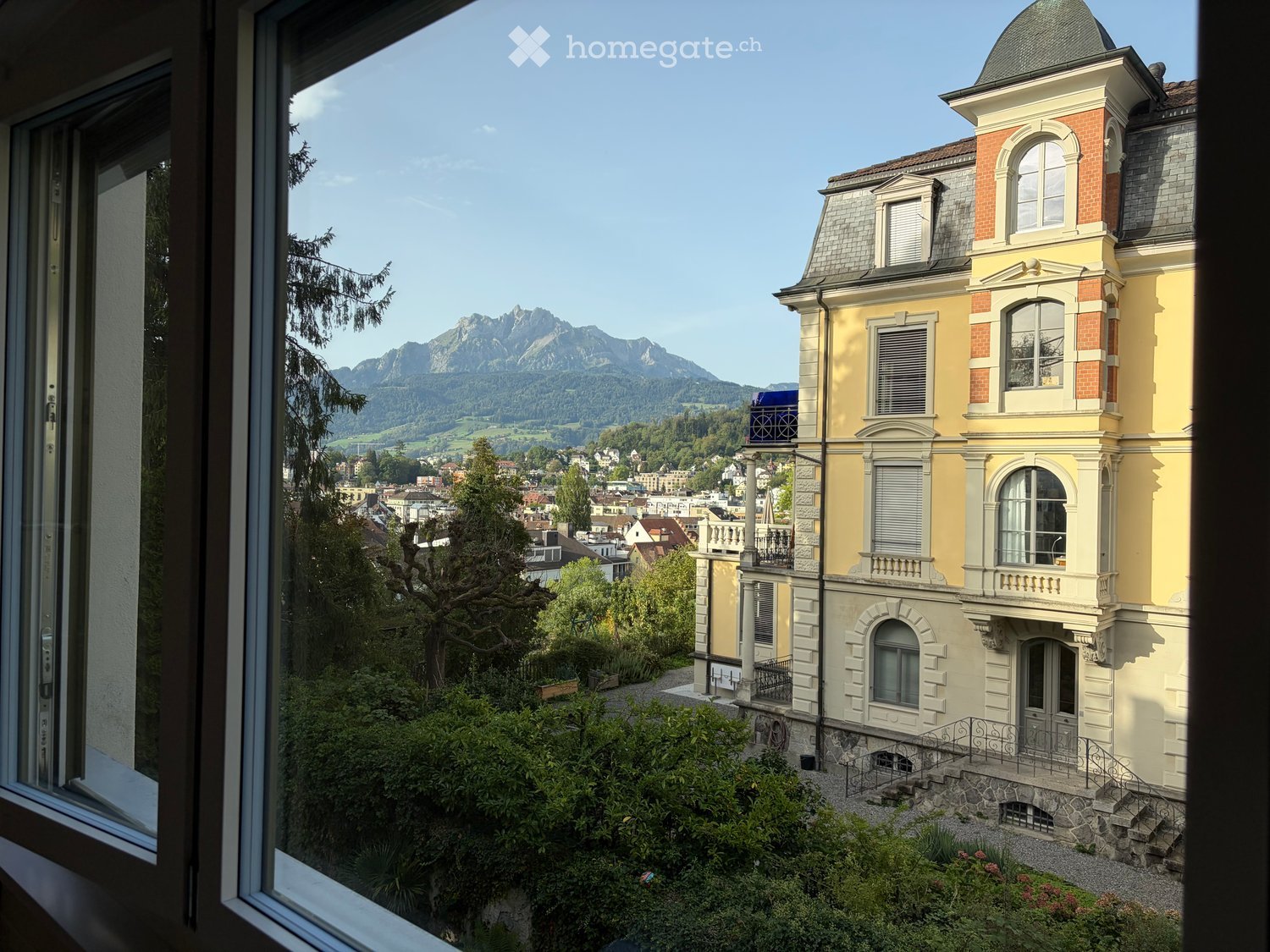 View from a window showing a large, yellow and white building with multiple stories and a dome-shaped roof, surrounded by greenery and trees, with a scenic mountain view in the background.