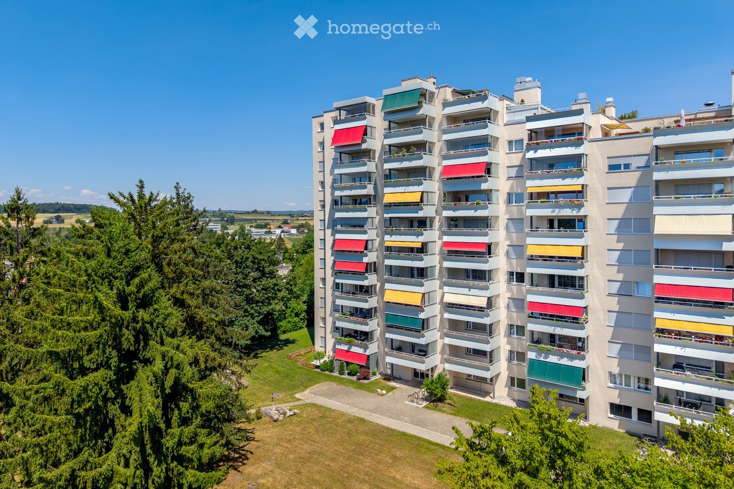 A modern multi-story apartment building with balconies, colorful awnings, and surrounding greenery.