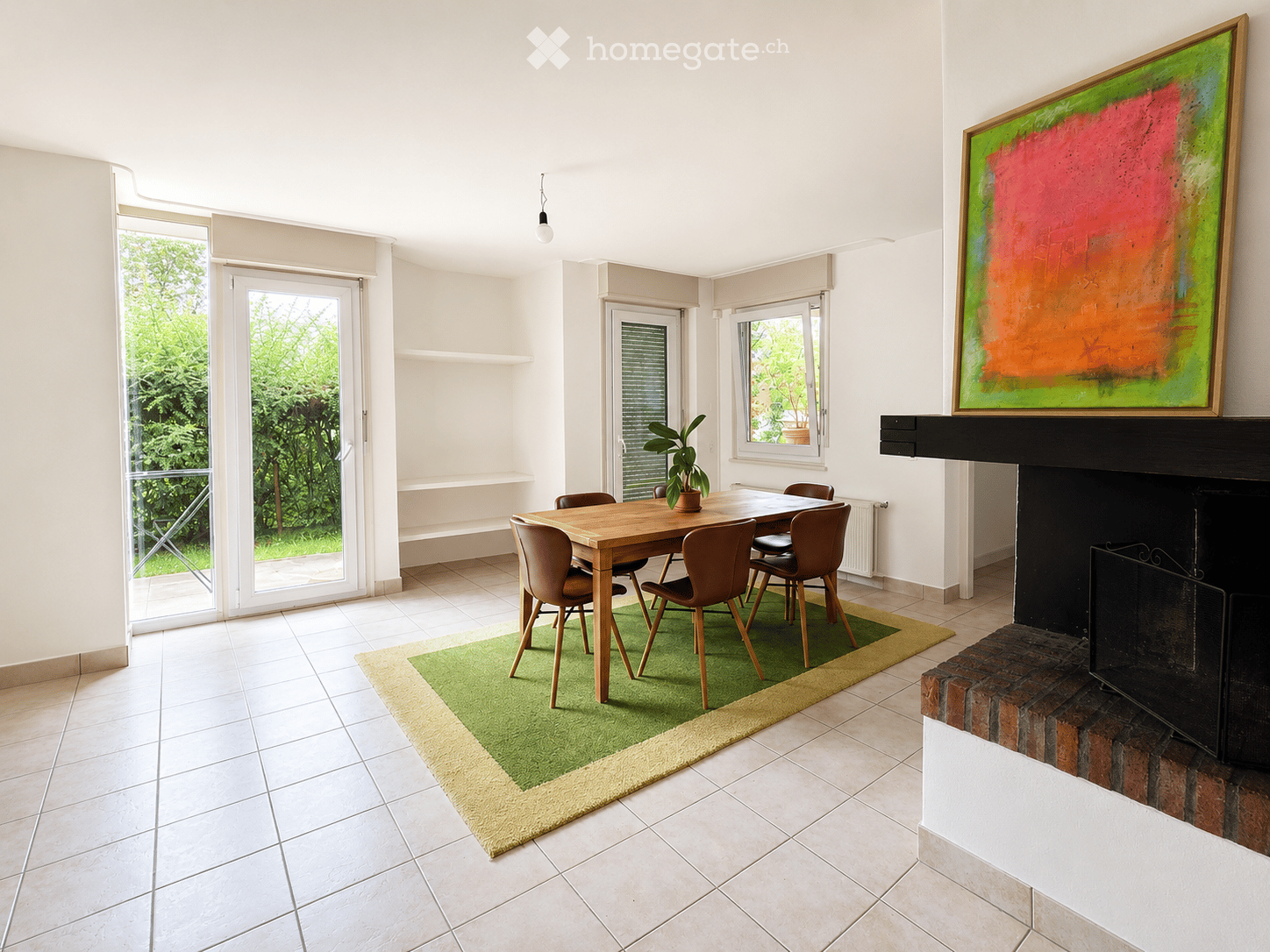 Bright dining room with a wooden dining table, six chairs, a green area rug, a fireplace with a painting above it, and a potted plant on the table. Large windows and sliding glass doors lead to a garden.