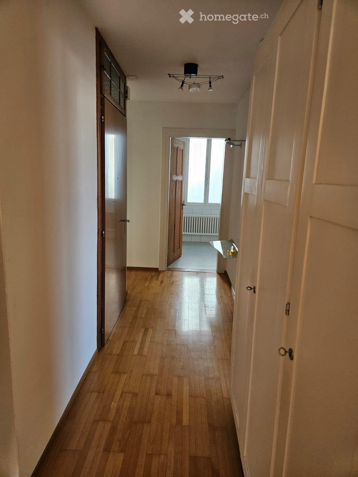 Empty hallway with wooden flooring, white walls, and several doors. A chandelier hangs from the ceiling, and a small shelf is visible on the right side.
