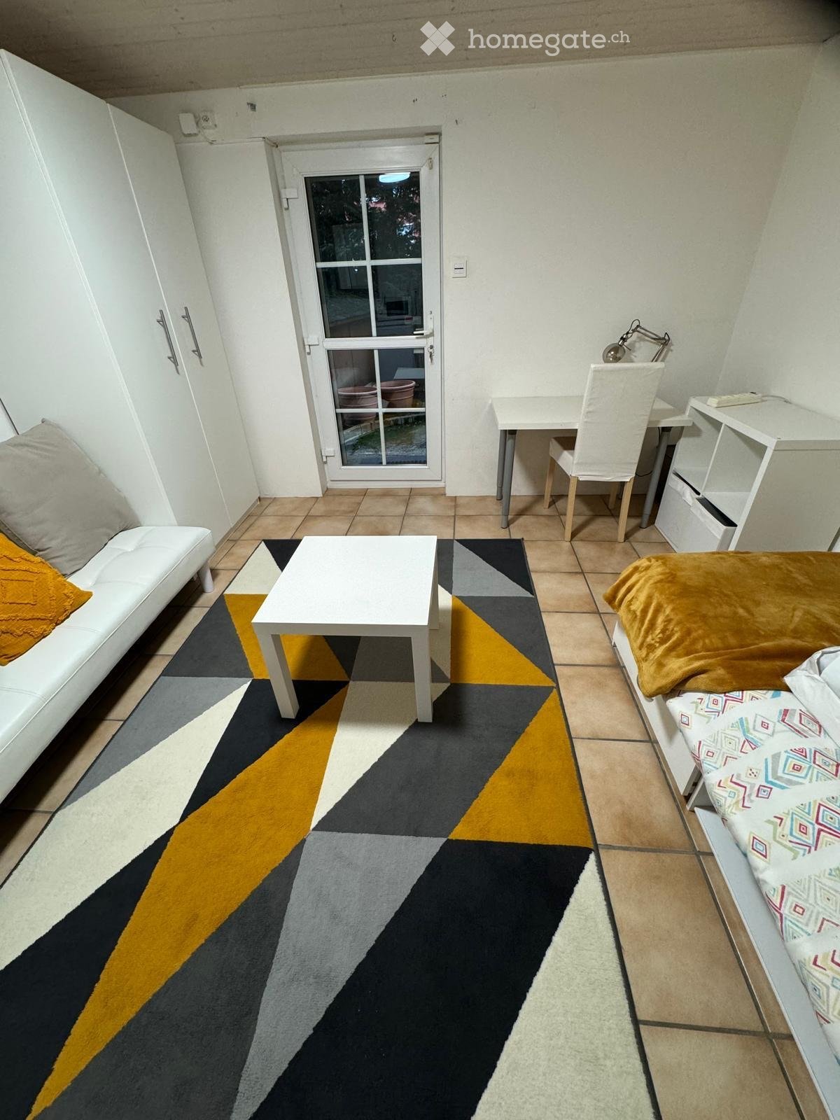 A compact, well-furnished room with a sofa, coffee table, bed, desk, and chair. The room features a geometric patterned rug and a sliding glass door leading outside.