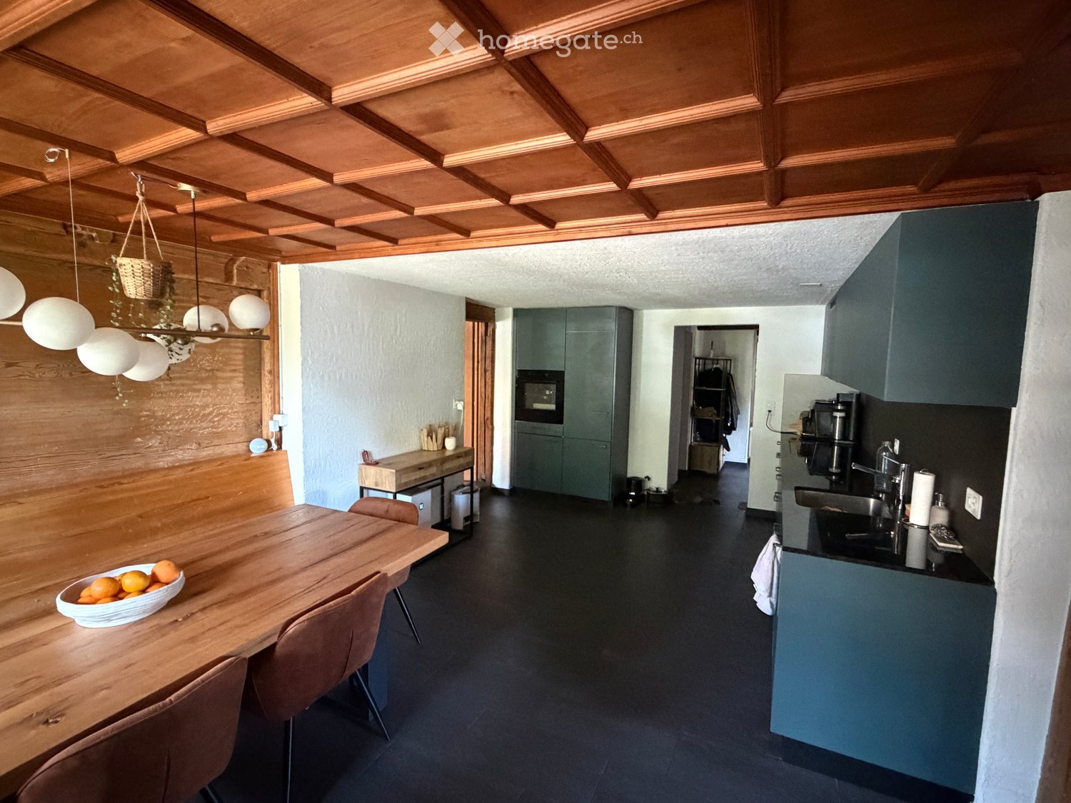 Modern kitchen with wooden ceiling, dark flooring, and grey cabinets. Features a wooden dining table with chairs, a bowl of oranges, and a hanging light fixture.