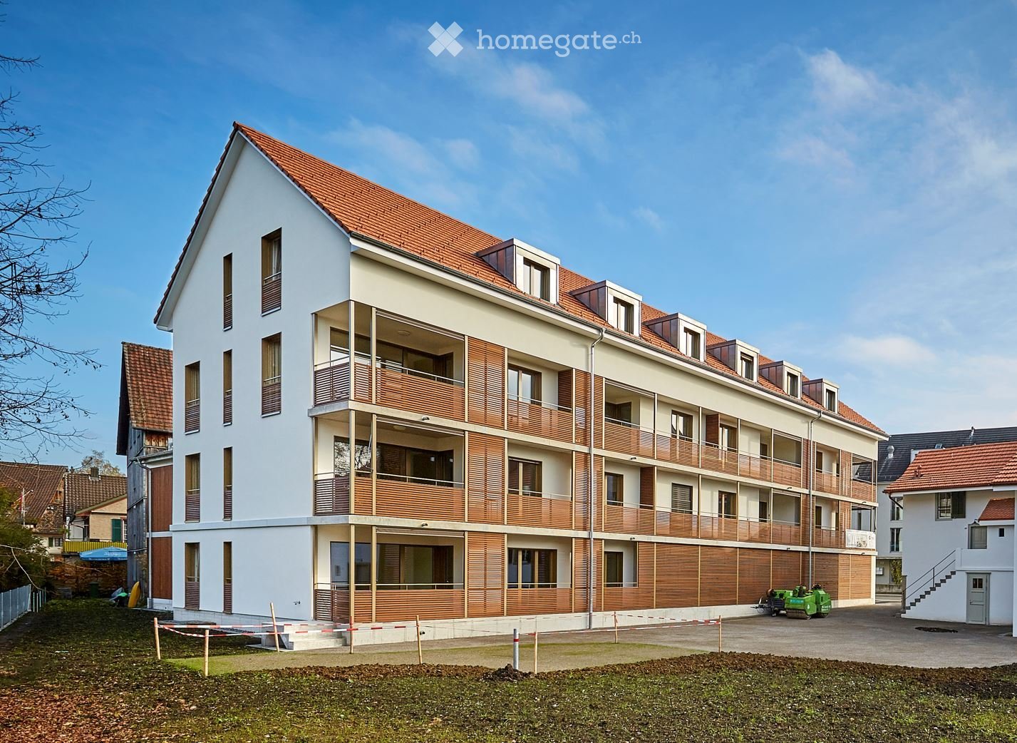 Modern multi-story residential building with white walls, wooden balconies, and red-tiled roof. The building features multiple windows and a raised ground floor entrance.