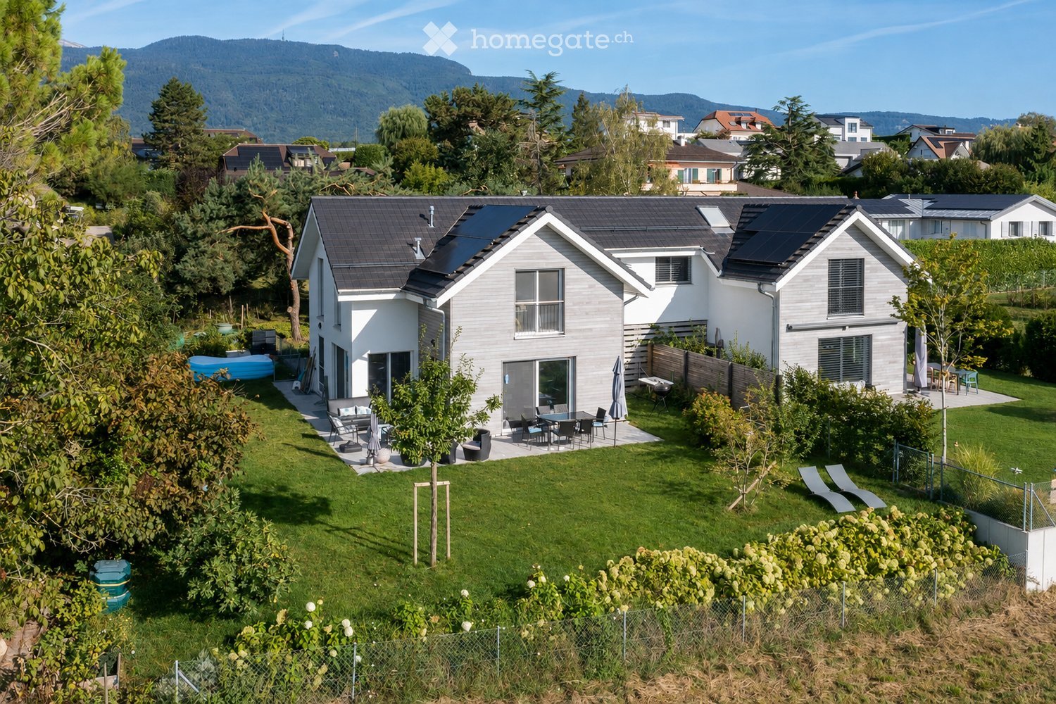 A modern house with a well-maintained garden, patio furniture, and solar panels on the roof, surrounded by trees and a scenic mountain backdrop.