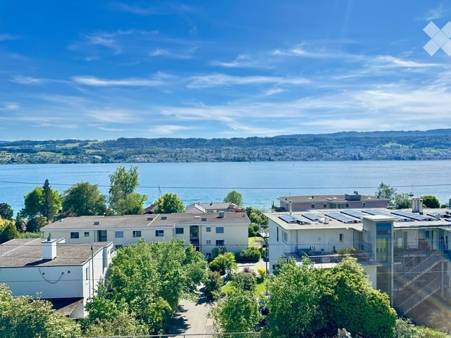 Aerial view of a residential complex with multiple buildings, surrounded by trees, overlooking a large body of water and distant hills.