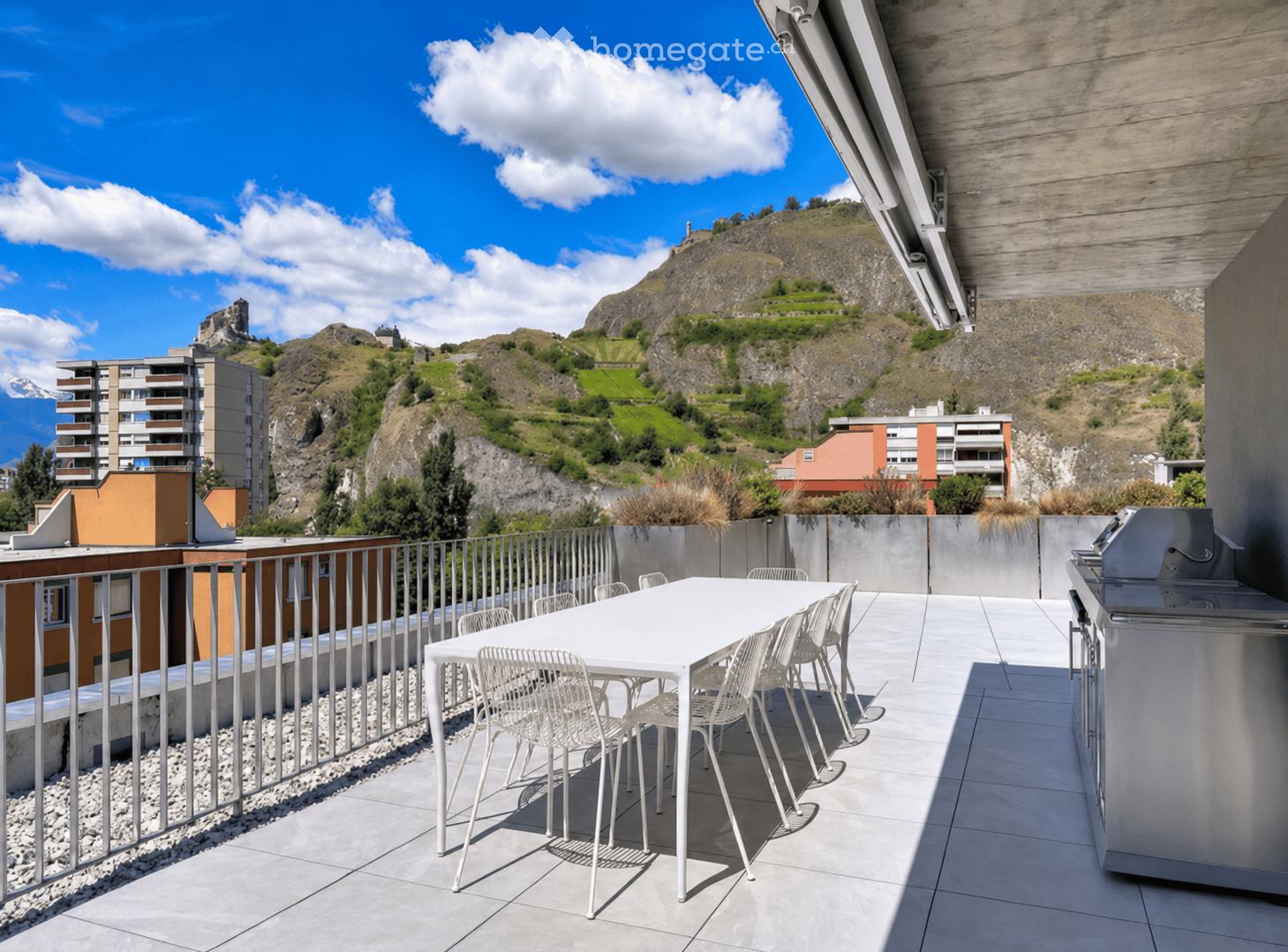 Modern rooftop terrace with a white dining table, chairs, and a grill, overlooking a scenic mountain view.