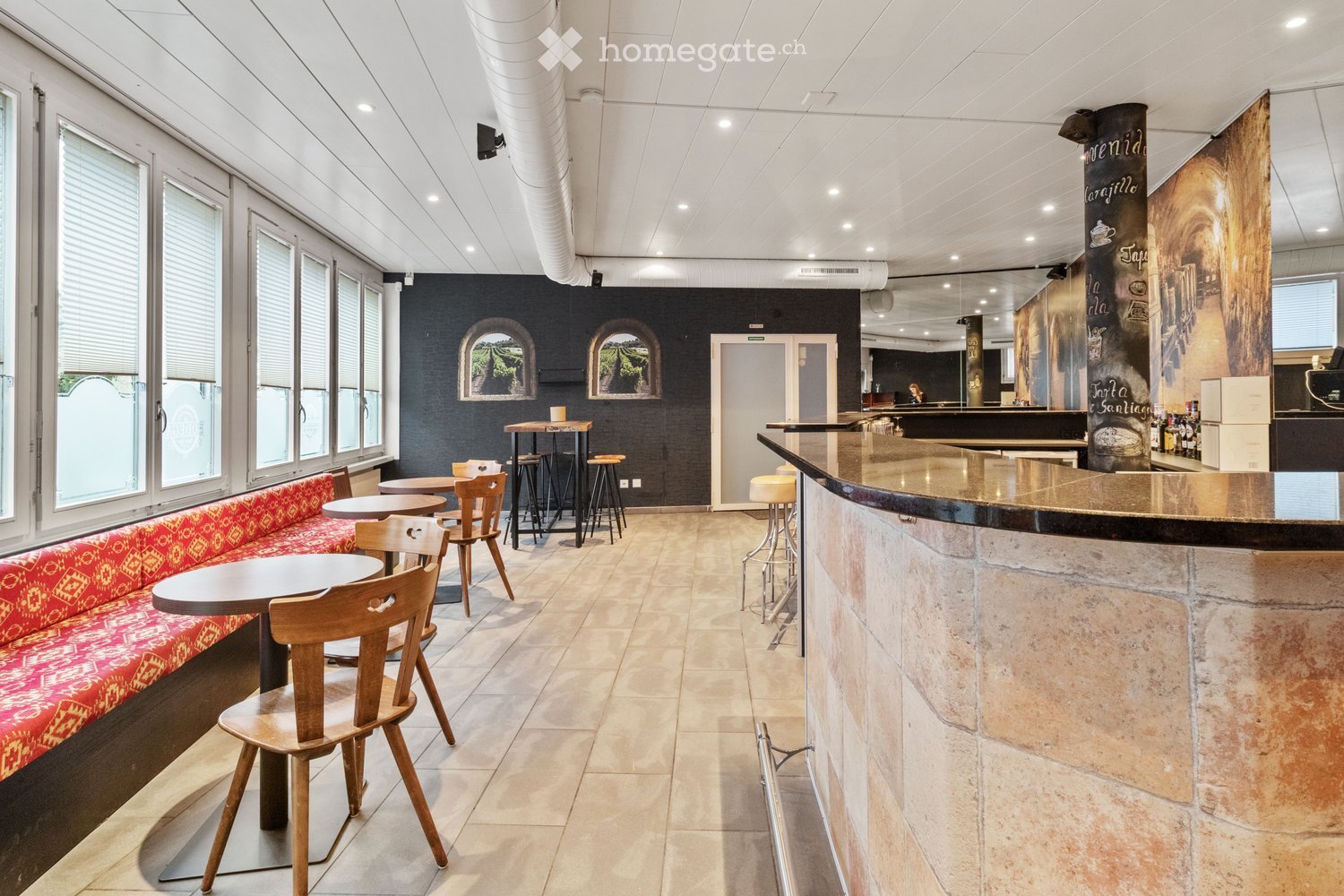 Modern interior with a mix of seating and bar area, featuring a red patterned sofa, wooden tables, chairs, and bar stools.