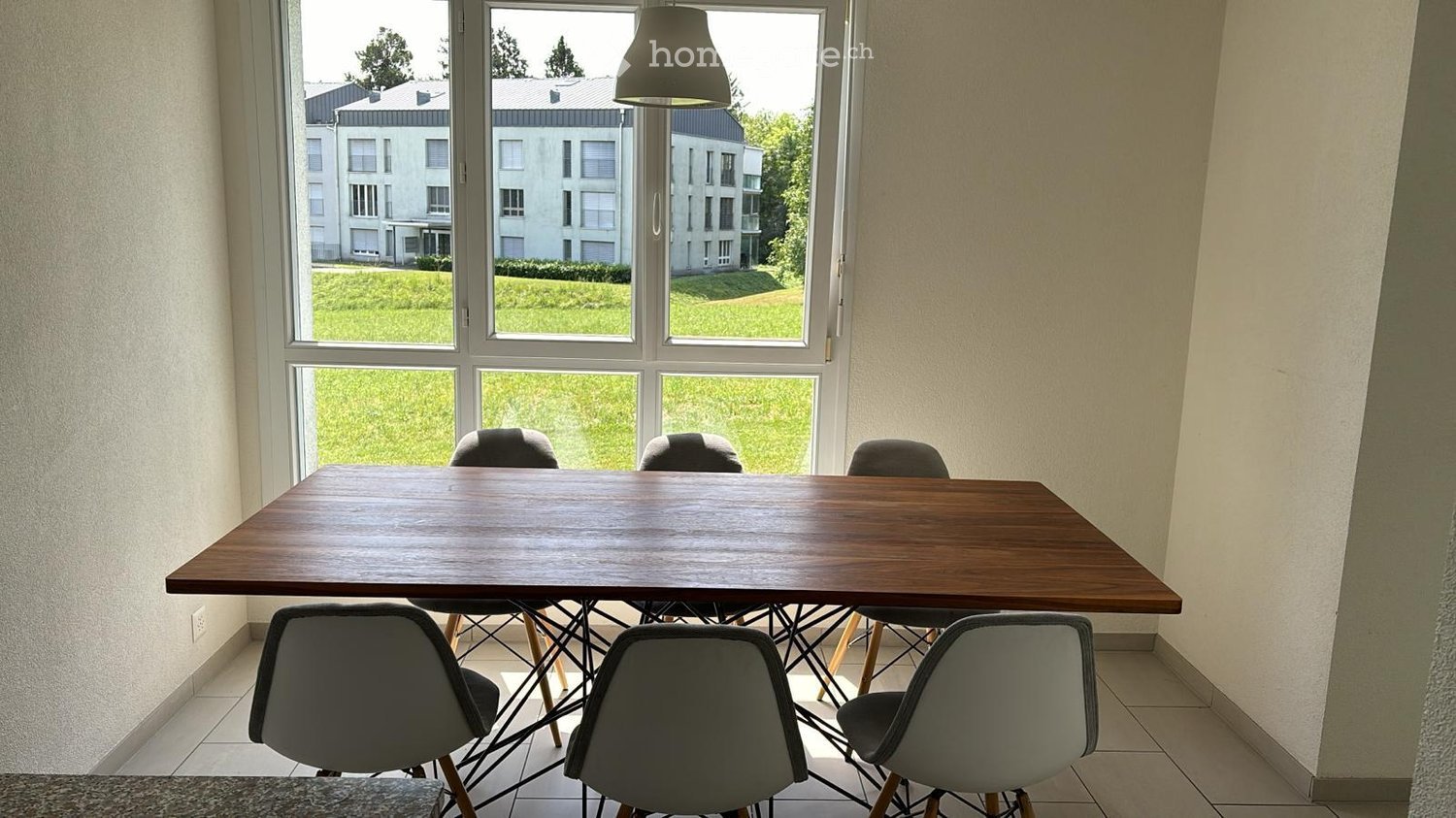 Bright room with large windows overlooking a green lawn and modern buildings. A wooden dining table is surrounded by six grey chairs.