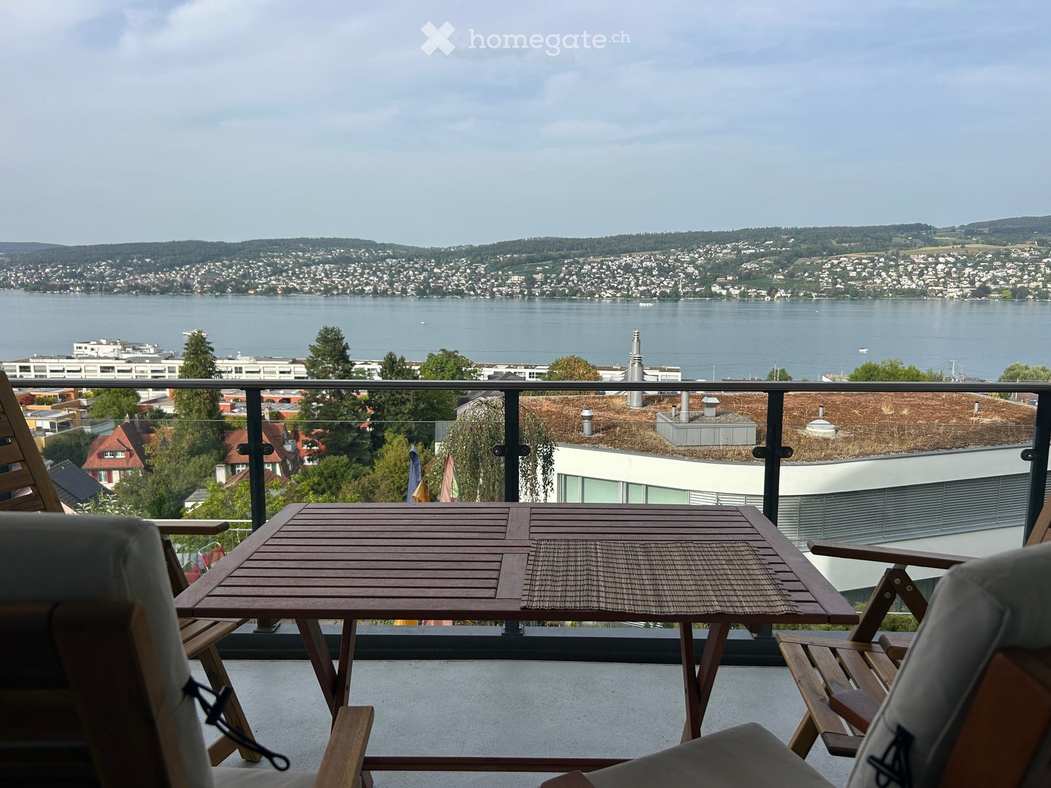 Balcony with wooden table and chairs, overlooking a scenic lake and cityscape.
