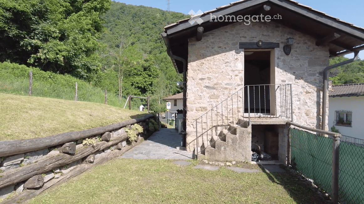 A rustic stone building with a small balcony and metal railing, surrounded by greenery and a wooden fence.