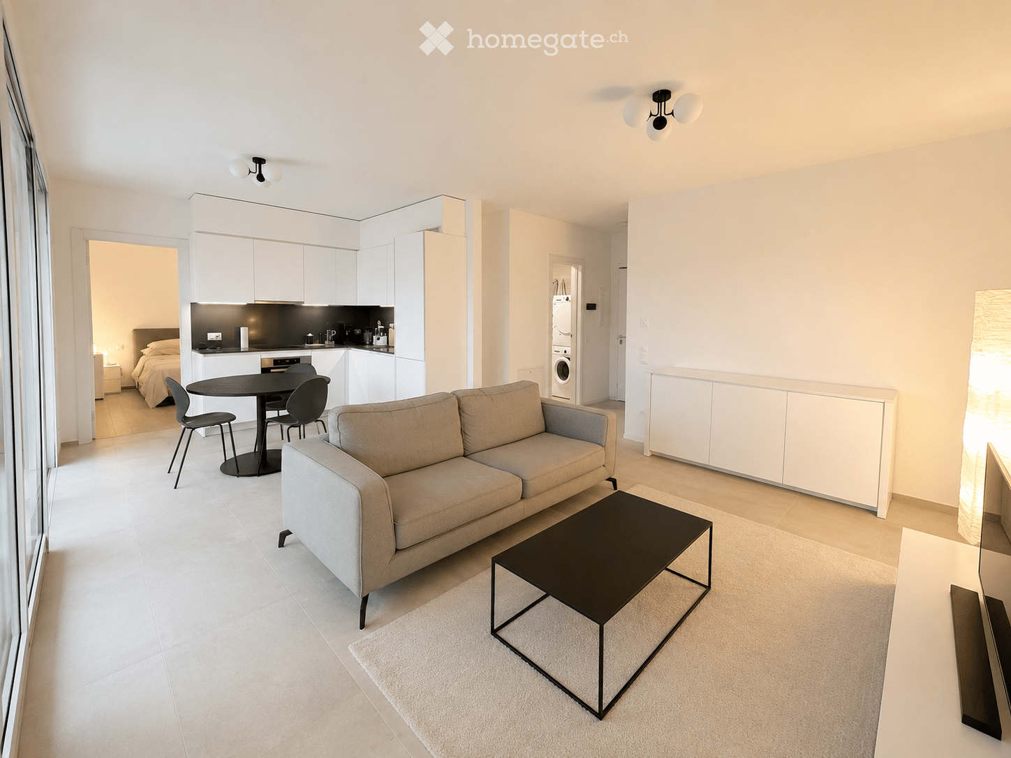 Modern, open-concept living space with a beige sofa, black coffee table, dining area with a round table and chairs, and a compact kitchen with white cabinets and stainless steel appliances. A bedroom area is visible in the background with a bed and nights