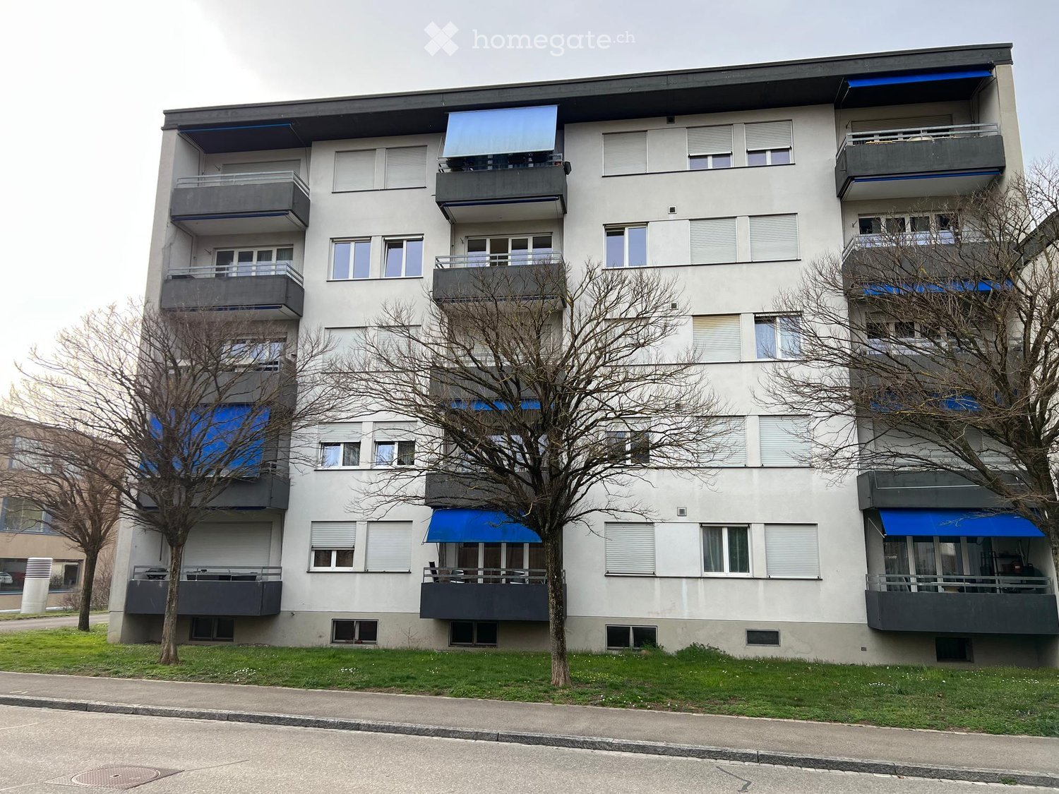 Four-story residential building with multiple balconies, large windows, and blue awnings. The building has a modern design with a mix of grey and white exterior walls.