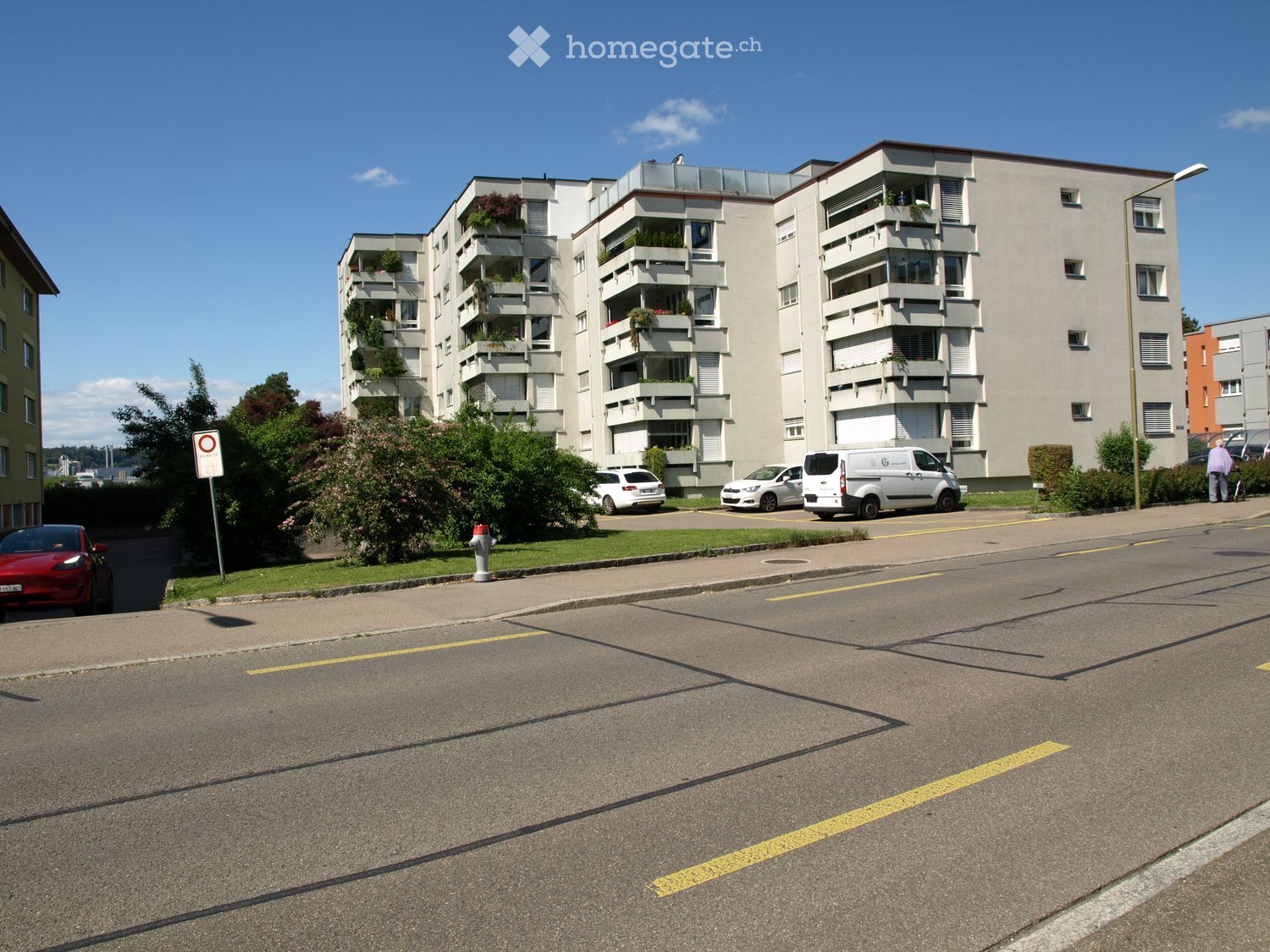 Multi-story residential building with balconies, parked cars, and greenery in front.
