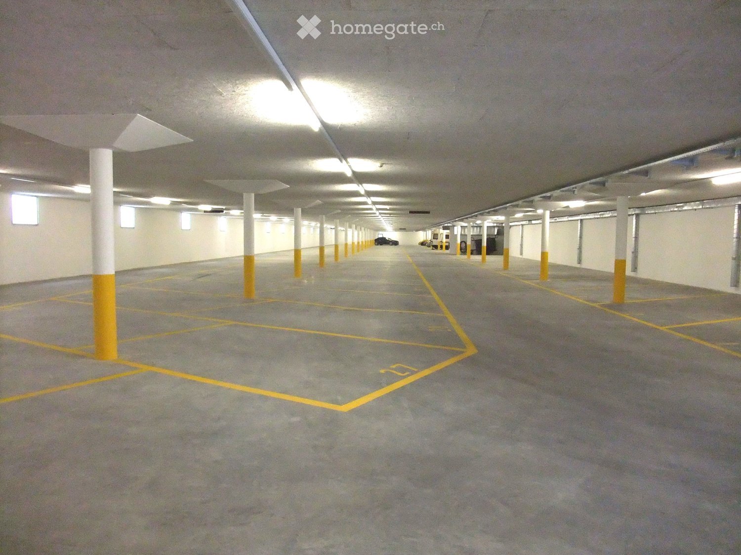 Large parking area with multiple vehicles parked, gray flooring, white and yellow painted pillars, windows on the ceiling, yellow parking lines