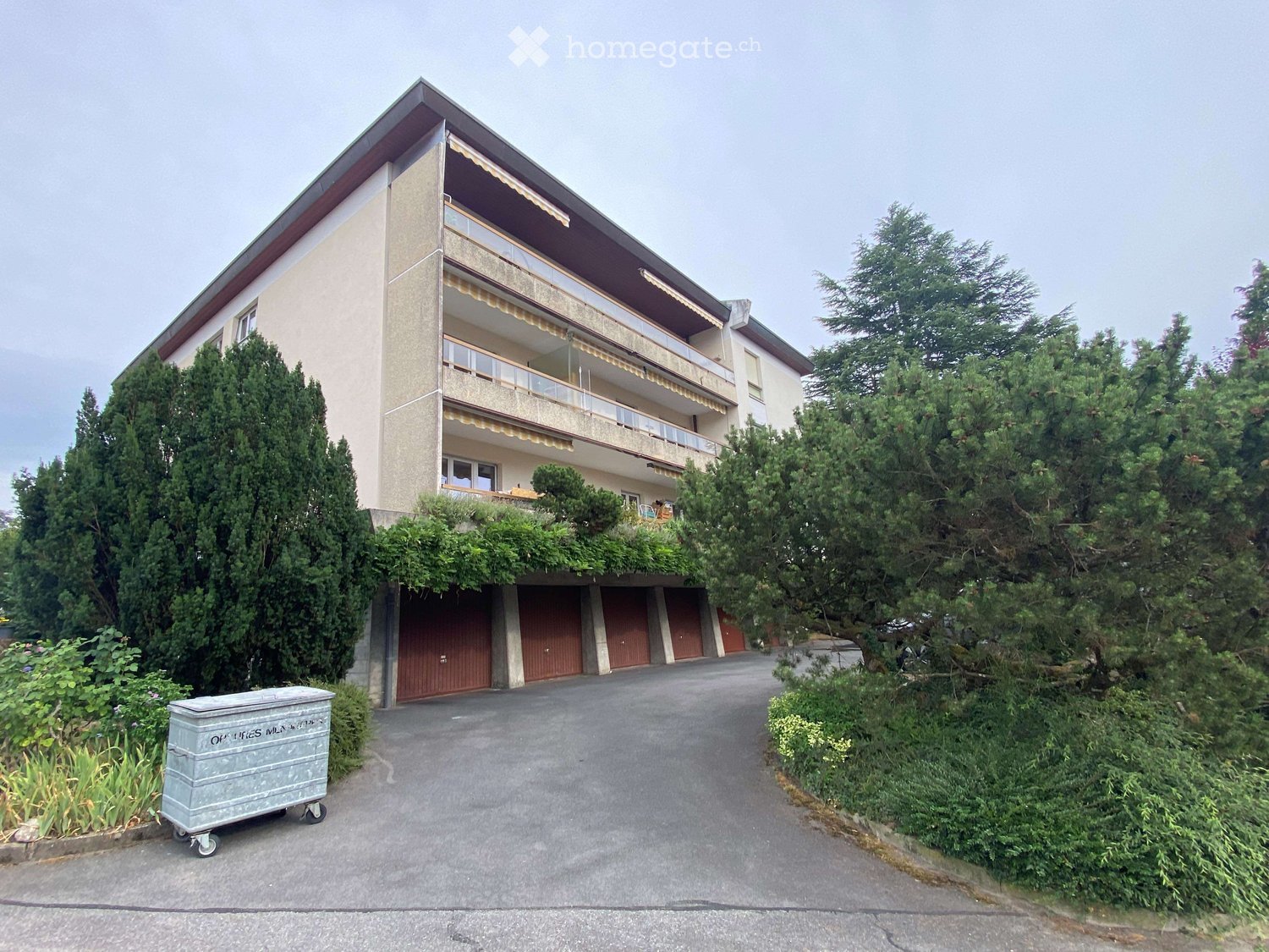 Two-story building with a balcony, multiple garage doors, and surrounding greenery.