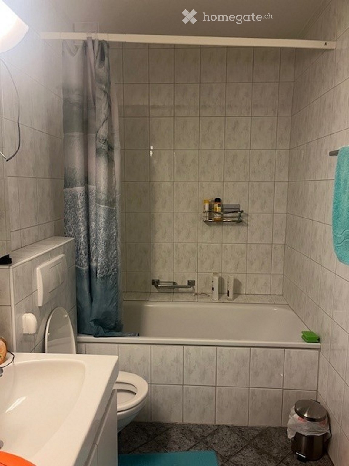 Bathroom with beige tiled walls and floor, featuring a bathtub, toilet, sink, shower curtain, towel, bathroom shelf, and a trash can.