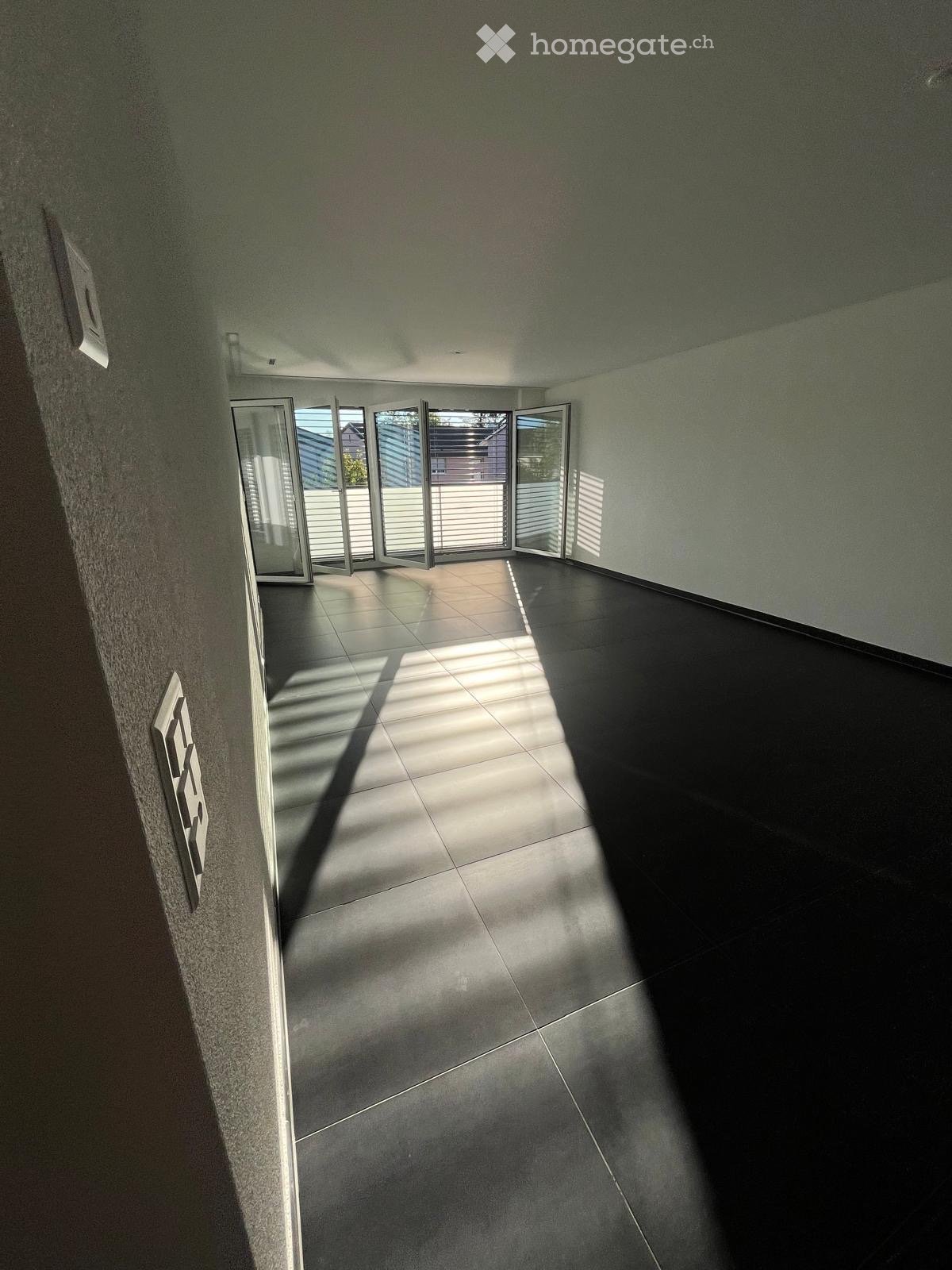 Empty hallway with tiled floor, large glass doors leading outside, and modern light fixtures on the walls.