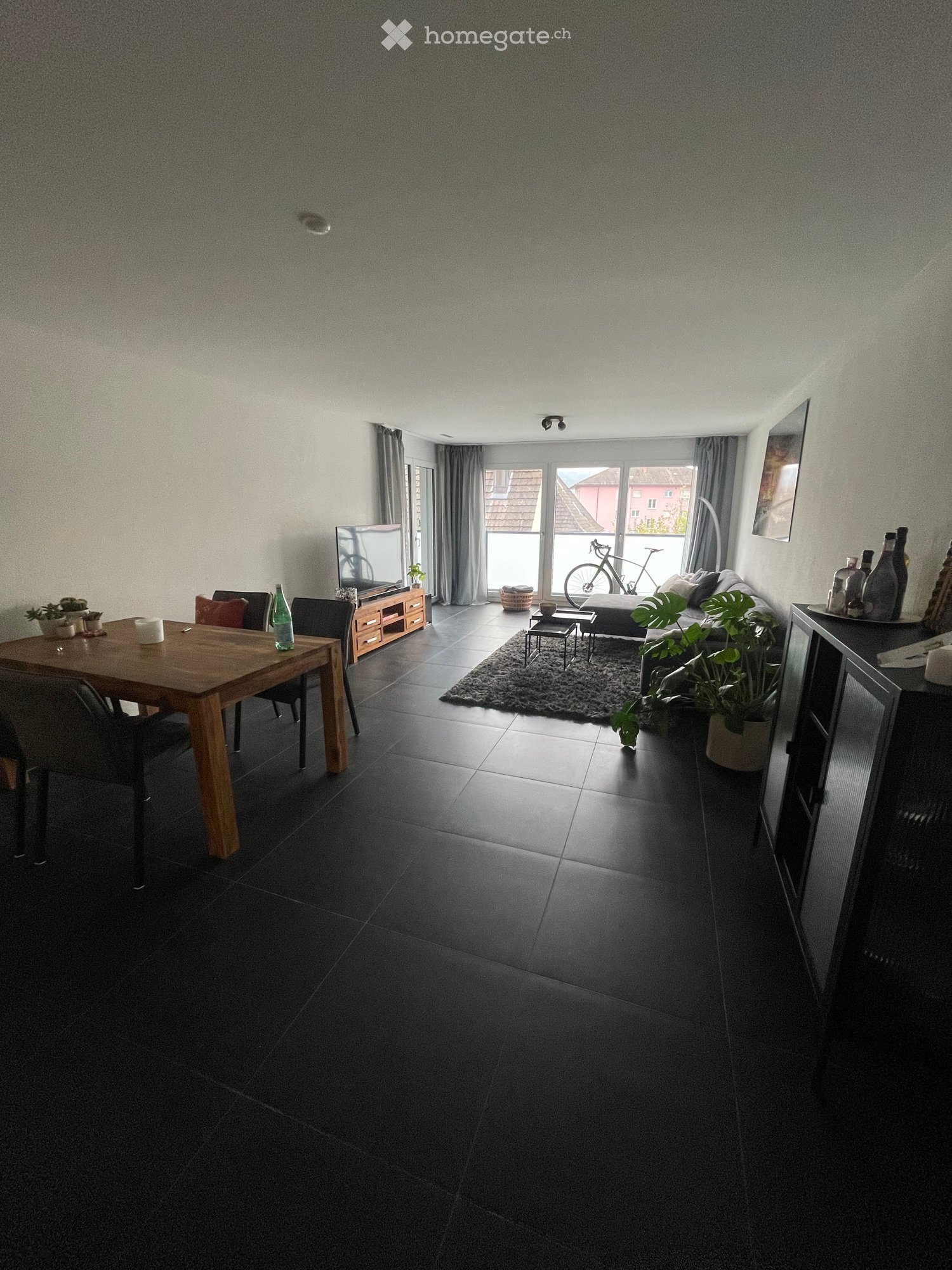 A modern, open-plan living and dining area with dark tiled flooring, a large sofa, a wooden dining table with chairs, a television on a stand, and several potted plants. The room features large windows with curtains and a view of a bicycle outside.