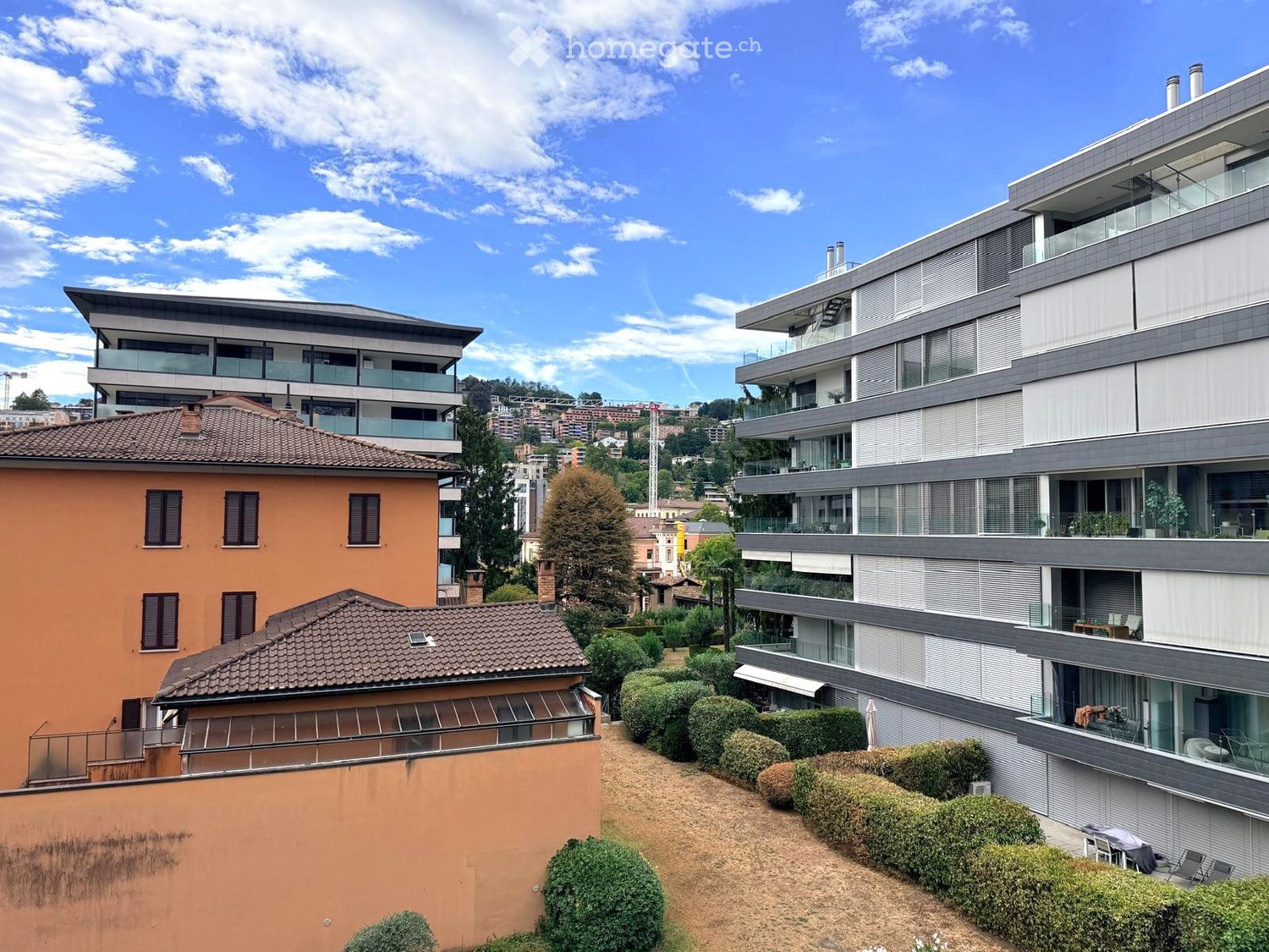 Modern multi-story residential building with balconies, surrounded by greenery and other structures in the background.