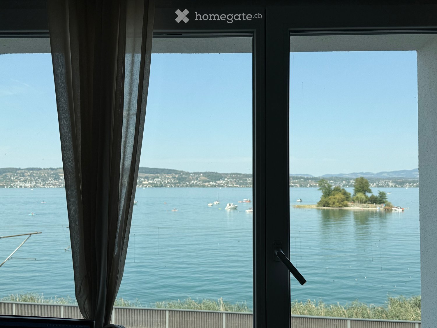 View from a window showing a lake with boats, distant hills, and a small island.