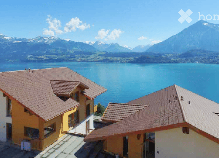 Building. Two-story residential building with a brown tiled roof and yellow walls, situated near a lake with mountain views in the background.