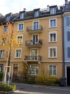 Multi-story residential building with balconies, windows, and a small garden area in front.
