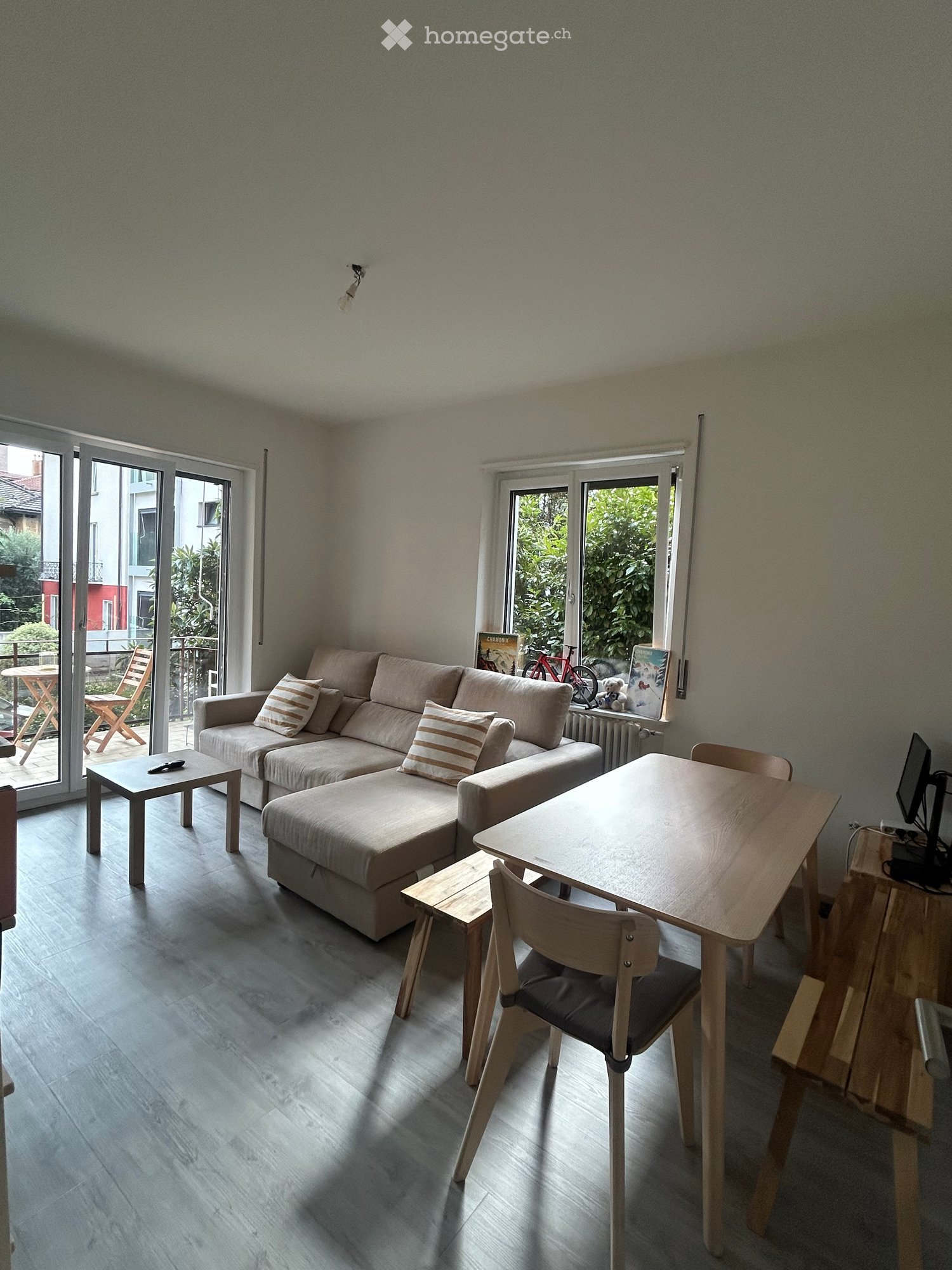 Modern living room with a grey sectional sofa, wooden coffee table, dining table with chairs, and a TV screen. Large windows and sliding doors lead to a balcony.